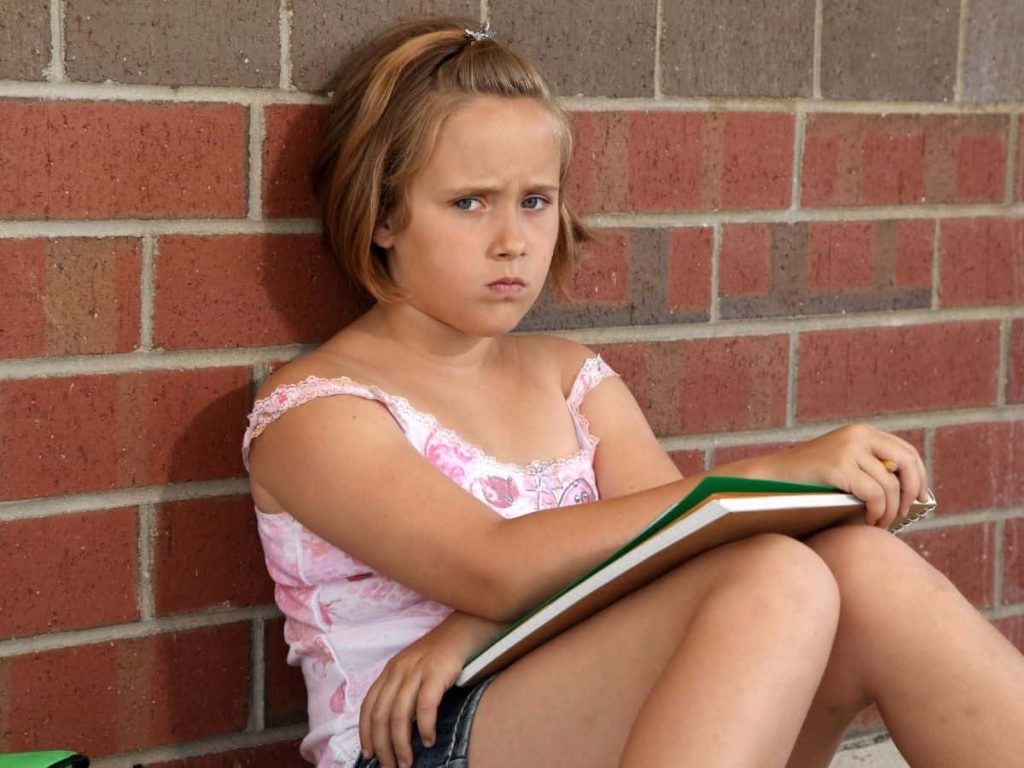 frowning girl with notebook in her lap leaning against brick wall